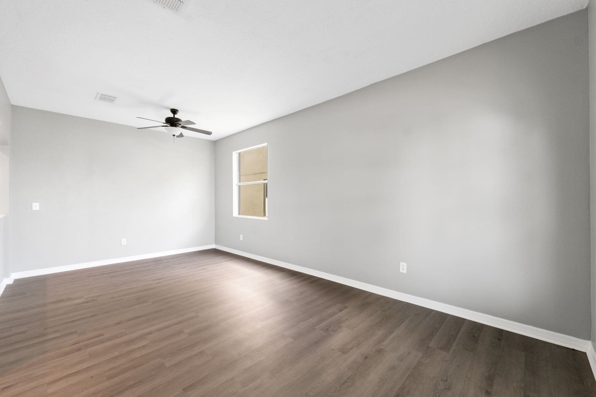 Empty bedroom with a ceiling fan, light wood floors and a window looking onto greenery.