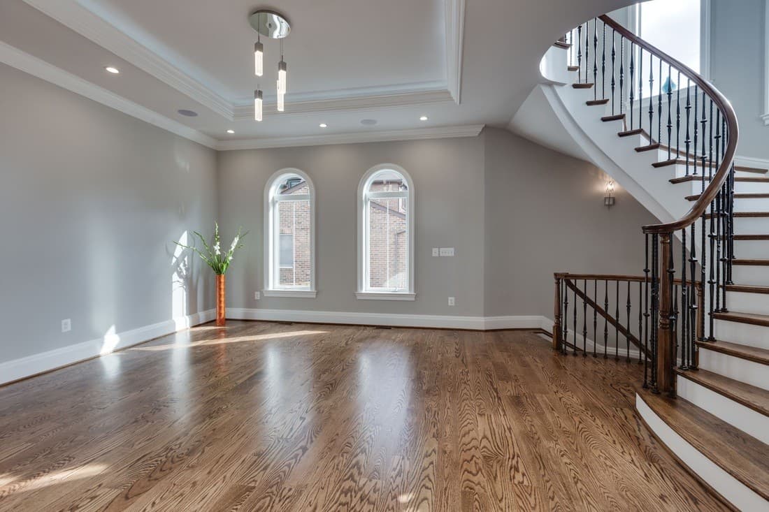 Empty formal living room with pale wood floors, tall windows and a curved staircase with a dark handrail on the right.