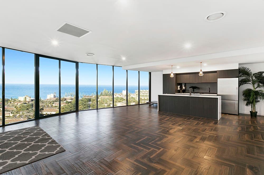 Empty open-plan living room with herringbone wood floors, a white kitchen along the back wall and floor-to-ceiling windows onto an ocean view.